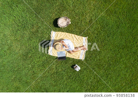Woman in a white dress and hat, sitting on the plaid on the green grass having a picnic with food basket. Top view, drone, aerial view Woman in a white dress and hat, sitting on the plaid on the green grass having a picnic with food basket. Top view, drone, aerial view 93858138