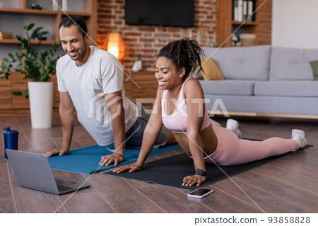 Glad young black couple in sportswear do stretching or practicing yoga, watch video lesson on laptop on mat Glad young black couple in sportswear do stretching or practicing yoga, watch video lesson on laptop on mat 93858828