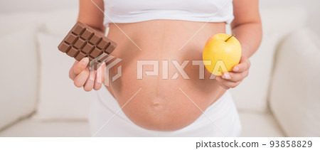 A pregnant woman is holding an apple and a bar of chocolate. The ninth month of pregnancy. 93858829