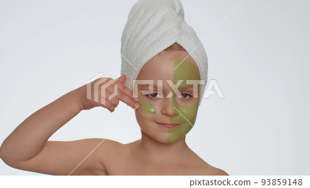 Beautiful smiling child girl kid in towel on head applying cleansing moisturizing green facial mask 93859148