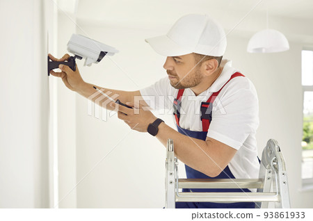 Electrician worker installs and screws surveillance video camera on wall in bright office space. 93861933