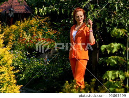 Charming red young woman in an orange trouser suit among flowers and greenery 93863399