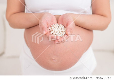 A faceless pregnant woman is holding a handful of white pills. 93870932