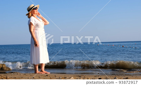 mature woman in a straw hat and white dress enjoying the sun walking along the blue sea coast on a mature woman in a straw hat and white dress enjoying the sun walking along the blue sea coast on a 93872917