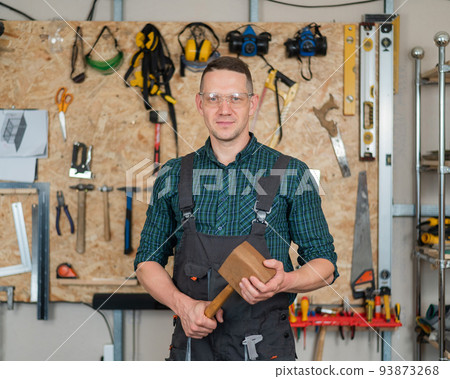 Portrait of a carpenter in goggles and overalls holding a wooden hammer in the workshop against the background of a wall with tools. Portrait of a carpenter in goggles and overalls holding a wooden hammer in the workshop against the background of a wall with tools. 93873268