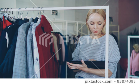 Young businesswoman is checking garments on rails and working with tablet in her clothing shop. She is checking prices and labels, touching screen and typing. Successful start-up concept. 93874395