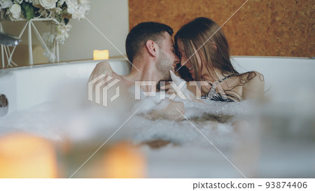 Adorable cheerful young couple is having fun in bubbling hot tub playing with foam, talking and kissing. Romantic relationships, happy people and wellness concept. 93874406