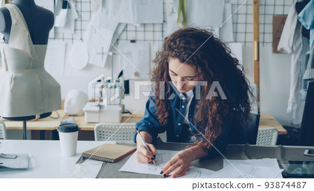 Young female artist fashion designer is drawing women's garment sketch with pencil at table in modern studio. Mannequin, sewing items, take-away coffee are visible. Young female artist fashion designer is drawing women's garment sketch with pencil at table in modern studio. Mannequin, sewing items, take-away coffee are visible. 93874487
