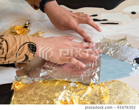 Female hands apply gold and silver potal to the picture. Potal painting technique, applying gold leaf particles to an interior painting. Female hands apply gold and silver potal to the picture. Potal painting technique, applying gold leaf particles to an interior painting. 93876302
