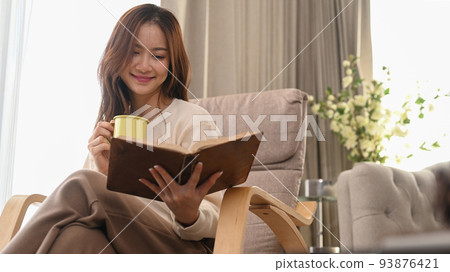 Pleasant young woman drinking herbal tea and reading book on armchair, spending leisure weekend at home 93876421