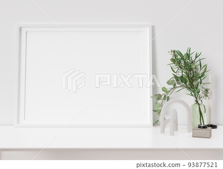 Empty horizontal picture frame standing on white shelf. Frame mock up. Copy space for picture, poster. Template for your artwork. Close up view. Plant in vase, home accessories, sculpture. 3D render. 93877521