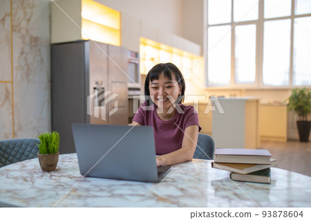 A girl in violet tshirt sitting at the laptop and looking happy A girl in violet tshirt sitting at the laptop and looking happy 93878604