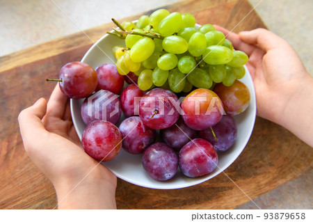 women's hands with a plate full of green grapes and plums on a wooden board, the concept of fresh fruit and healthy food 93879658