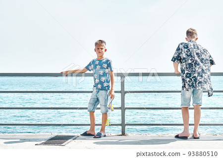 Schoolboys wearing casual clothes stand at pier handrail. Couple of young brothers enjoys spending summer holidays and travelling together at seaside Schoolboys wearing casual clothes stand at pier handrail. Couple of young brothers enjoys spending summer holidays and travelling together at seaside 93880510