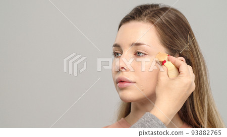Makeup artist or stylist applies foundation using wet sponge to the face of the female model. Visagiste  applying cosmetic concealer base on a face. 93882765