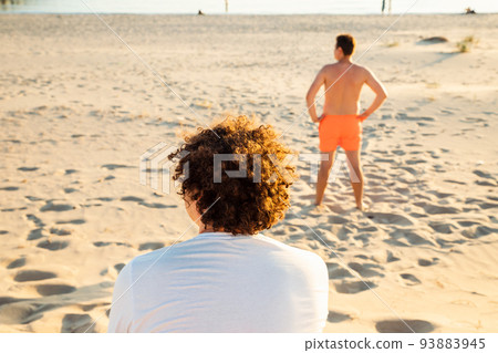 Back turned look away two young men on sandy summer beach. Curly hair man is sitting, another man is lifeguard in red swim shorts. 93883945