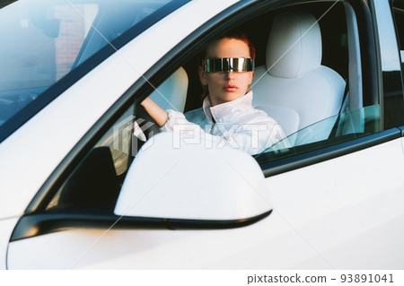 Portrait of fashion cyberpunk girl driving a white supercar. Red hair woman with futuristic eyeglasses sits by the car steering wheel. Future technology concept. Selective focus, copy space. Portrait of fashion cyberpunk girl driving a white supercar. Red hair woman with futuristic eyeglasses sits by the car steering wheel. Future technology concept. Selective focus, copy space. 93891041