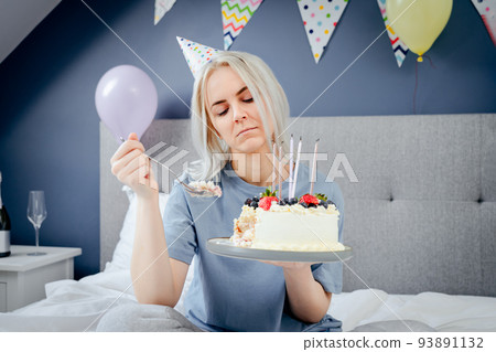 Sad, upset woman in pajama and party cap thinking eat or not to eat birthday cake sitting on the bed in decorated bedroom. Celebrates birthday alone concept. Selective focus Sad, upset woman in pajama and party cap thinking eat or not to eat birthday cake sitting on the bed in decorated bedroom. Celebrates birthday alone concept. Selective focus 93891132