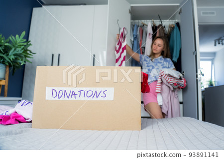 A cardboard box of selected clothes for donating to a Charity shop standing on the bed. Decluttering, Sorting clothes, and Cleaning Up. Reuse, second-hand. Conscious consumer, sustainability. 93891141