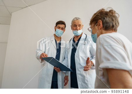 Doctors in masks explain to a sick patient in a wheelchair her diagnosis and treatment 93891148