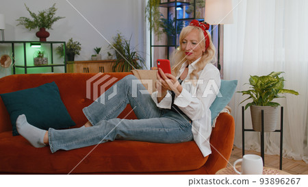 Senior old grandmother sitting on sofa, using smartphone share messages on social media application 93896267