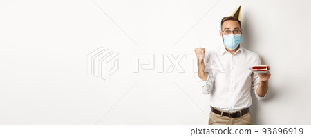 Covid-19, social distancing and celebration. Hopeful happy birthday man in face mask, holding bday cake and rejoicing, standing against white background 93896919