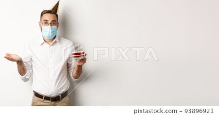 Coronavirus, quarantine and holidays. Confused man in face mask, holding birthday cake and shrugging, standing over white background clueless 93896921