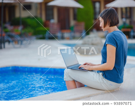 Young business woman working at computer by hotel pool. 93897071