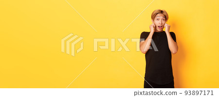 Excited handsome blond guy in black clothes, looking tempted and standing over yellow background 93897171