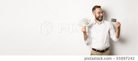 Excited businessman holding money and looking at credit card, standing over white background Excited businessman holding money and looking at credit card, standing over white background 93897242