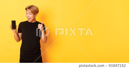 Portrait of satisfied asian guy looking at smartphone screen and showing thumbs-up in approval, standing yellow background 93897282