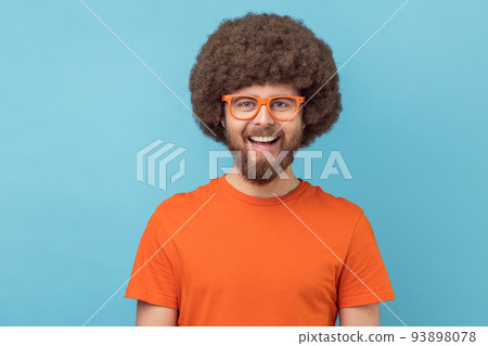 Portrait of funny positive man with Afro hairstyle wearing orange T-shirt and eyeglasses, looking at camera with happy expression and toothy smile. Indoor studio shot isolated on blue background. 93898078