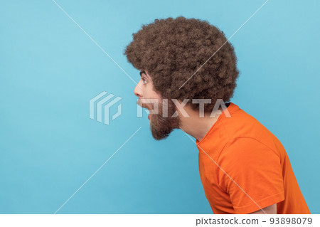 Side view of amazed man with Afro hairstyle wearing orange T-shirt standing with mouth open in surprise, has shocked expression, hears unbelievable news. Indoor studio shot isolated on blue background 93898079