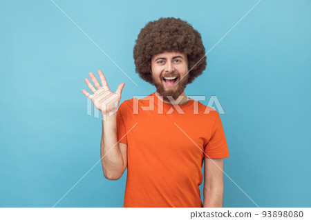 Portrait of cheerful friendly man with Afro hairstyle wearing orange T-shirt saying hi and waving hand, greeting, looking at camera. Indoor studio shot isolated on blue background. 93898080