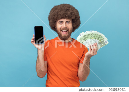 Portrait of satisfied man with Afro hairstyle wearing orange T-shirt showing euro banknotes and smart phone with empty display for promotional text. Indoor studio shot isolated on blue background. Portrait of satisfied man with Afro hairstyle wearing orange T-shirt showing euro banknotes and smart phone with empty display for promotional text. Indoor studio shot isolated on blue background. 93898081
