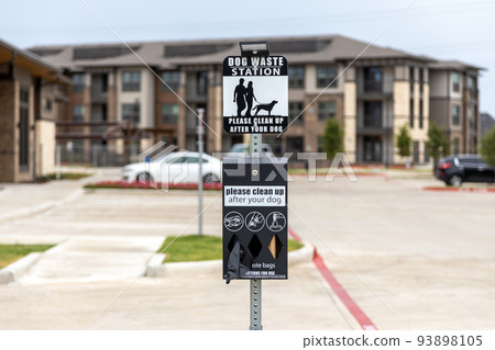 Dog waster station sign with plastic bags for waste. Clean after your dog while walking in residential complex, waste station with baggies with building and car parking on background Dog waster station sign with plastic bags for waste. Clean after your dog while walking in residential complex, waste station with baggies with building and car parking on background 93898105