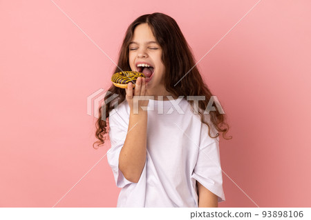 Sugar addiction. Portrait of satisfied little girl wearing white T-shirt biting delicious donut, looking with desire to eat sweet dessert. Indoor studio shot isolated on pink background. Sugar addiction. Portrait of satisfied little girl wearing white T-shirt biting delicious donut, looking with desire to eat sweet dessert. Indoor studio shot isolated on pink background. 93898106