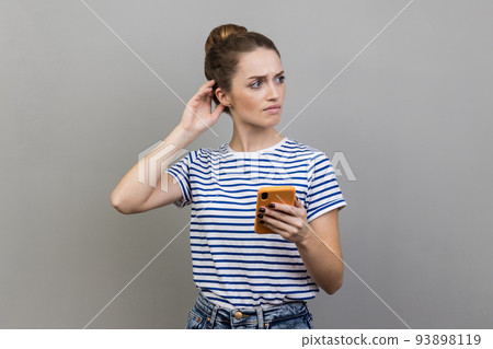 Portrait of pensive thoughtful woman wearing striped T-shirt standing with mobile phone, touching head, having doubt suspicion feeling. Indoor studio shot isolated on gray background. Portrait of pensive thoughtful woman wearing striped T-shirt standing with mobile phone, touching head, having doubt suspicion feeling. Indoor studio shot isolated on gray background. 93898119