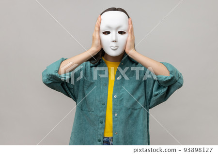 Portrait of unknown woman holding white mask, standing covering face, multiple personality disorder, wearing casual style jacket. Indoor studio shot isolated on gray background. 93898127