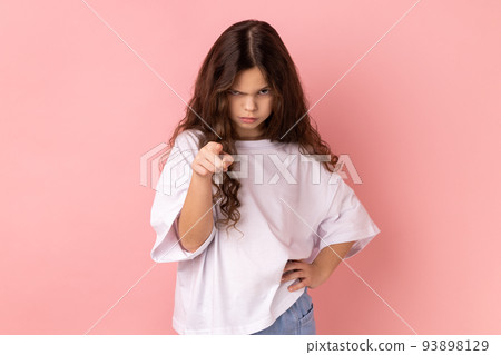 Portrait of angry little girl wearing white T-shirt noticing and pointing finger to camera, accusing with serious bossy face, making choice. Indoor studio shot isolated on pink background. 93898129