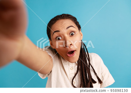 Amazed shocked woman with dreadlocks has surprised expression, making selfie, looking at camera with big eyes, point of view photo, wearing white shirt. Indoor studio shot isolated on blue background. Amazed shocked woman with dreadlocks has surprised expression, making selfie, looking at camera with big eyes, point of view photo, wearing white shirt. Indoor studio shot isolated on blue background. 93898130