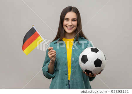 Smiling satisfied woman holding in hands flag of Germany and football ball, supporting favourite team on championship, wearing casual style jacket. Indoor studio shot isolated on gray background. Smiling satisfied woman holding in hands flag of Germany and football ball, supporting favourite team on championship, wearing casual style jacket. Indoor studio shot isolated on gray background. 93898131