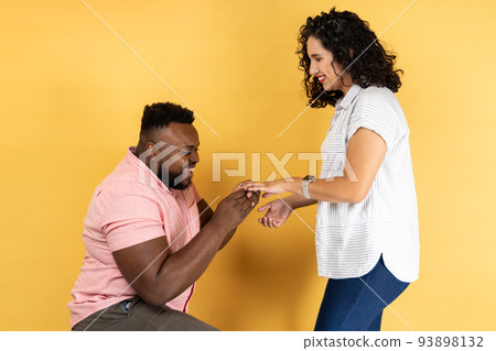 Portrait of happy lovely young couple in casual clothing standing together, romantic man puts on a wedding ring to her girlfriend, making proposal. Indoor studio shot isolated on yellow background. Portrait of happy lovely young couple in casual clothing standing together, romantic man puts on a wedding ring to her girlfriend, making proposal. Indoor studio shot isolated on yellow background. 93898132