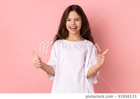Portrait of smiling little girl wearing white T-shirt showing thumbs up, like gesture, looking at camera demonstrating her satisfaction. Indoor studio shot isolated on pink background. Portrait of smiling little girl wearing white T-shirt showing thumbs up, like gesture, looking at camera demonstrating her satisfaction. Indoor studio shot isolated on pink background. 93898133