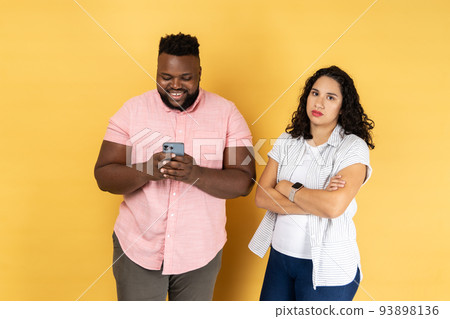 Satisfied young couple in casual clothing standing together, man using mobile phone and smiling, offended lonely woman standing with folded arms. Indoor studio shot isolated on yellow background. Satisfied young couple in casual clothing standing together, man using mobile phone and smiling, offended lonely woman standing with folded arms. Indoor studio shot isolated on yellow background. 93898136