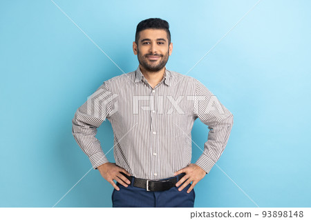 Portrait of happy attractive bearded businessman with smile holding hands on hips, looking at camera with confident expression, wearing striped shirt. Indoor studio shot isolated on blue background. 93898148