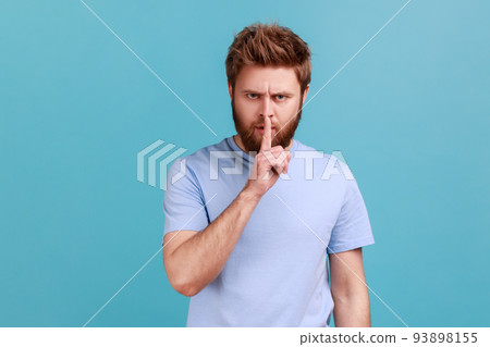 Portrait of bearded man holding finger near lips, showing shh gesture asking to keep silence and be quite, privacy, looking at camera. Indoor studio shot isolated on blue background. 93898155