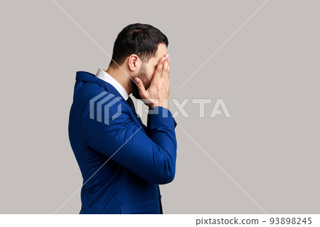 Side view of bearded man closing eyes with hand, dont want to see that, ignoring problems, hiding from stressful situations, wearing official style suit. Indoor studio shot isolated on gray background 93898245