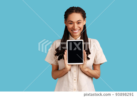 Happy positive woman holds modern tablet with mockup screen for your advertisement, poses with electronic gadget in hands, wearing white shirt. Indoor studio shot isolated on blue background. 93898291