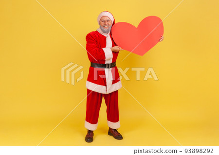 Full length portrait of elderly man with gray beard wearing santa claus costume holding big red heart, looking at camera with positive expression. Indoor studio shot isolated on yellow background. Full length portrait of elderly man with gray beard wearing santa claus costume holding big red heart, looking at camera with positive expression. Indoor studio shot isolated on yellow background. 93898292
